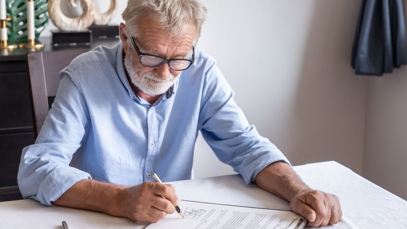 Senior man signing a document at home, focused and wearing glasses.