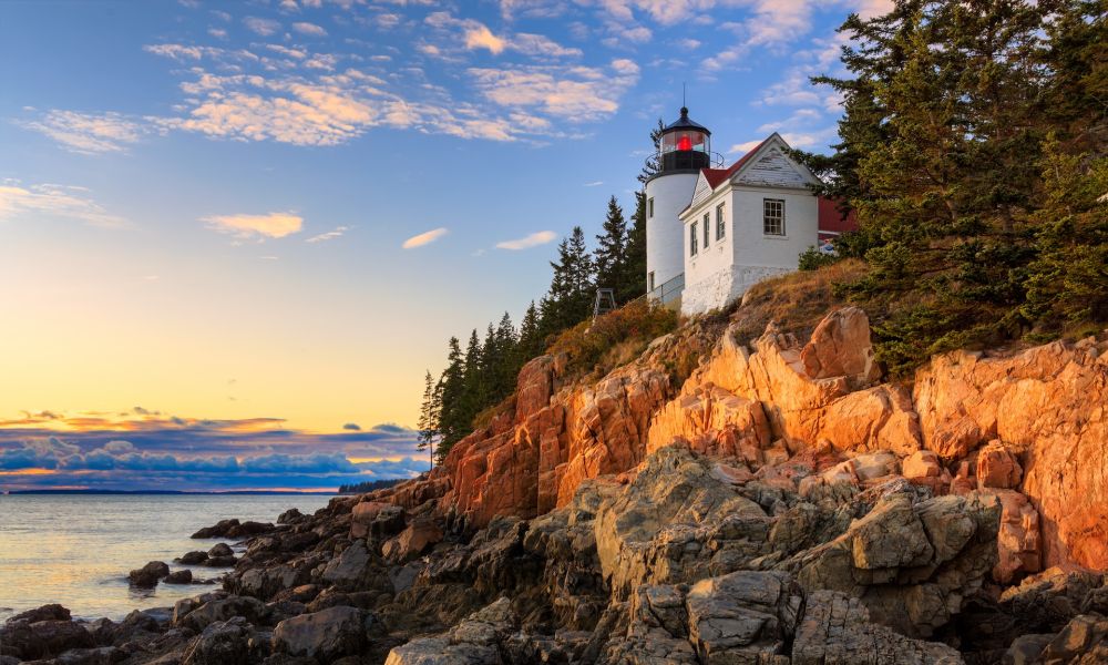 Lighthouse on a rocky cliff overlooking the ocean at sunset with a vibrant sky and pine trees.