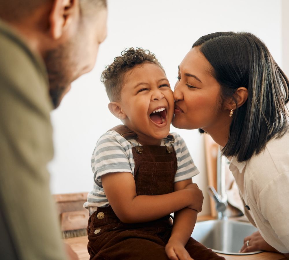 Mother kissing laughing child in kitchen, happy family bonding moment at home.