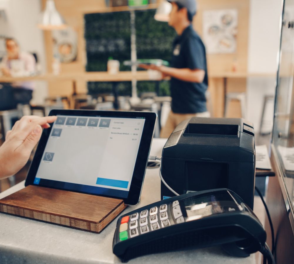 Tablet POS system in cafe with customer interaction, payment terminal, and food display in view.