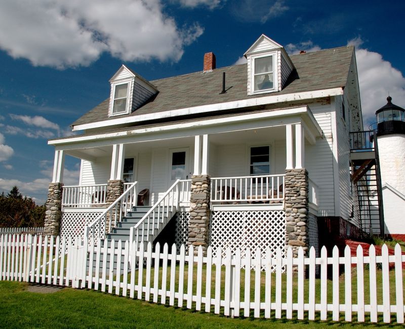 White house with stone pillars and picket fence near a lighthouse under a blue sky.