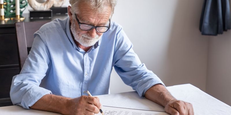 Senior man signing a document at home, focused and wearing glasses.