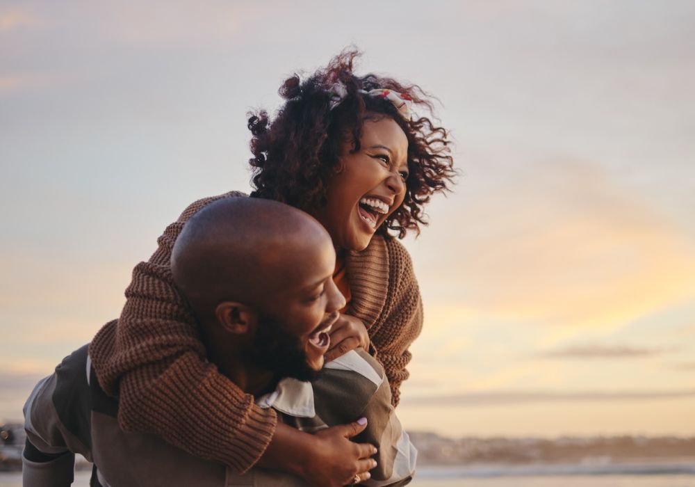 Happy couple enjoying a playful piggyback ride on the beach at sunset, laughing and smiling.