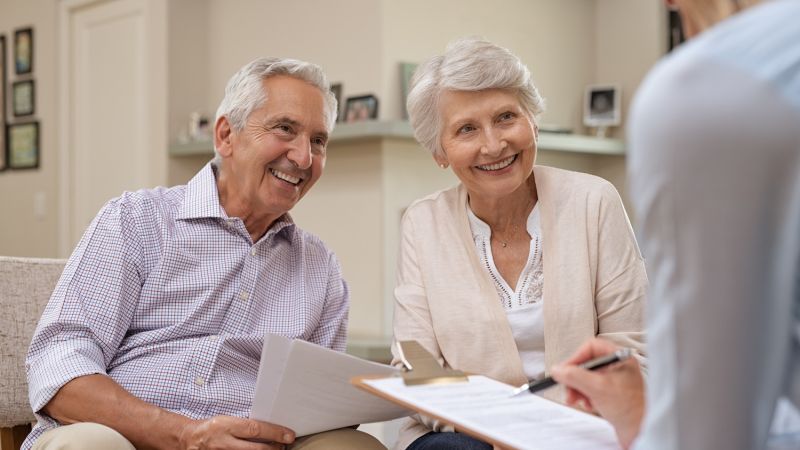 Senior couple discussing financial planning with advisor, smiling and holding documents in cozy living room setting.