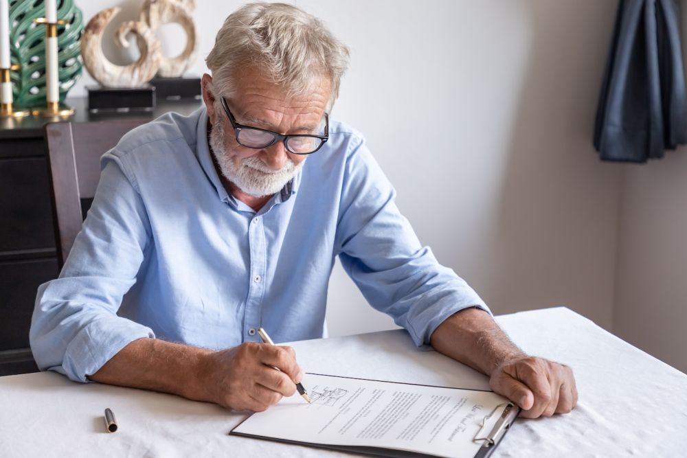Senior man signing a document at home, focused and wearing glasses.