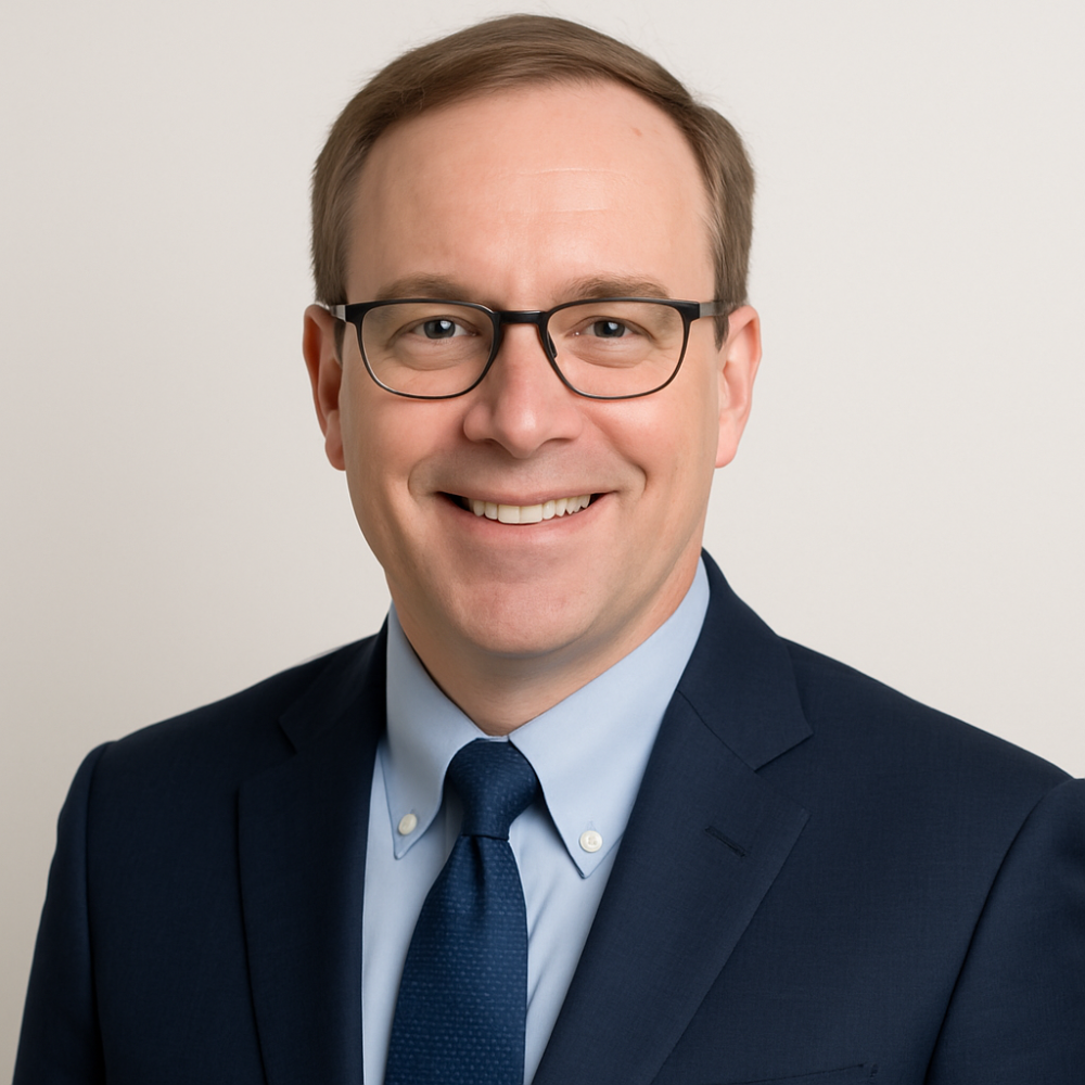 Smiling man in glasses wearing a suit and tie, professional portrait against a light background.