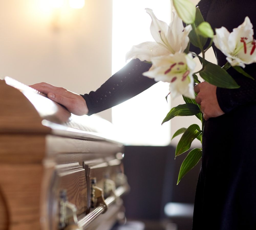 Person holding lilies, placing hand on wooden casket during funeral ceremony, expressing respect and condolences.