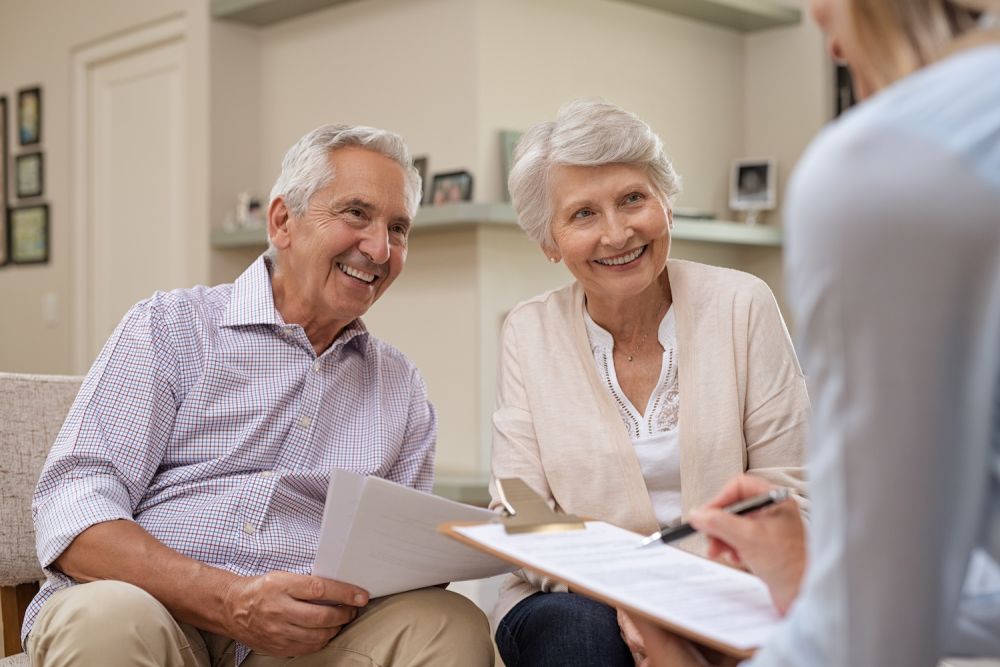 Senior couple discussing financial planning with advisor, smiling and holding documents in cozy living room setting.