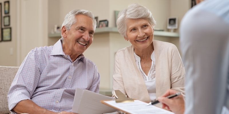Senior couple discussing financial planning with advisor, smiling and holding documents in cozy living room setting.