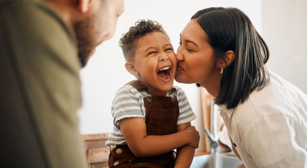 Mother kissing laughing child in kitchen, happy family bonding moment at home.