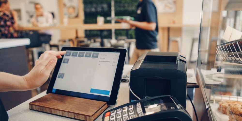 Tablet POS system in cafe with customer interaction, payment terminal, and food display in view.