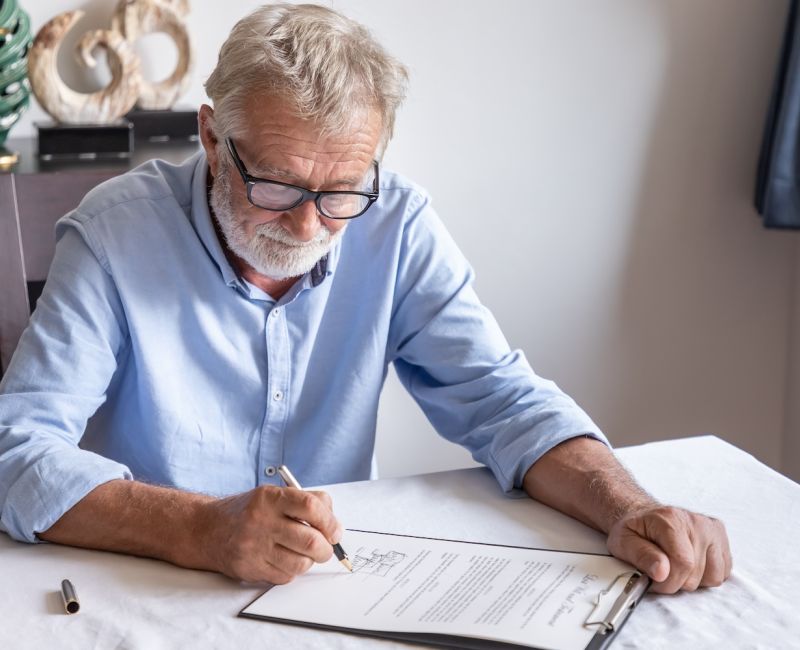 Senior man signing a document at home, focused and wearing glasses.