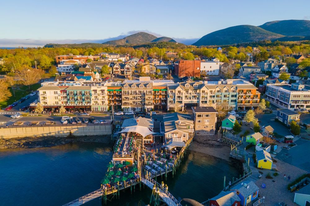 Aerial view of Bar Harbor waterfront in Maine, featuring colorful buildings and mountain backdrop under clear skies.