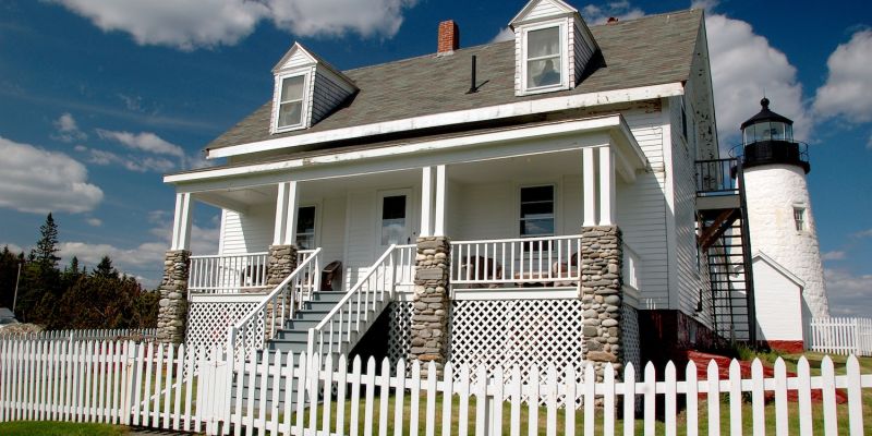 White house with stone pillars and picket fence near a lighthouse under a blue sky.