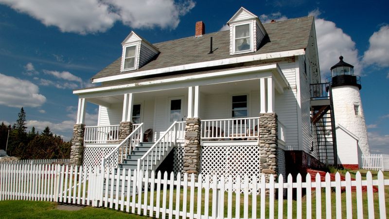 White house with stone pillars and picket fence near a lighthouse under a blue sky.