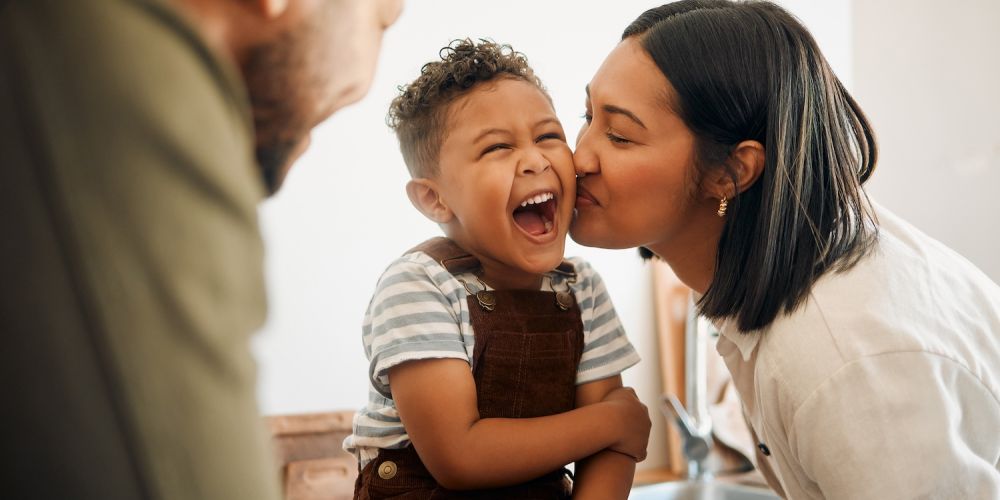 Mother kissing laughing child in kitchen, happy family bonding moment at home.
