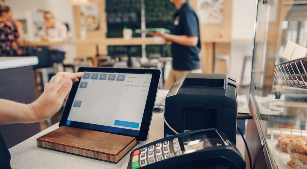 Tablet POS system in cafe with customer interaction, payment terminal, and food display in view.