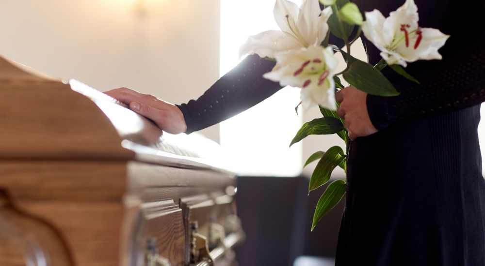 Person holding lilies, placing hand on wooden casket during funeral ceremony, expressing respect and condolences.