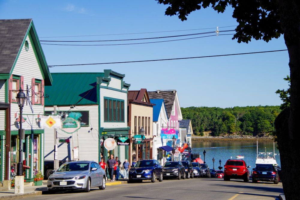 Charming coastal town street with colorful buildings, cars, and waterfront view under a clear blue sky.