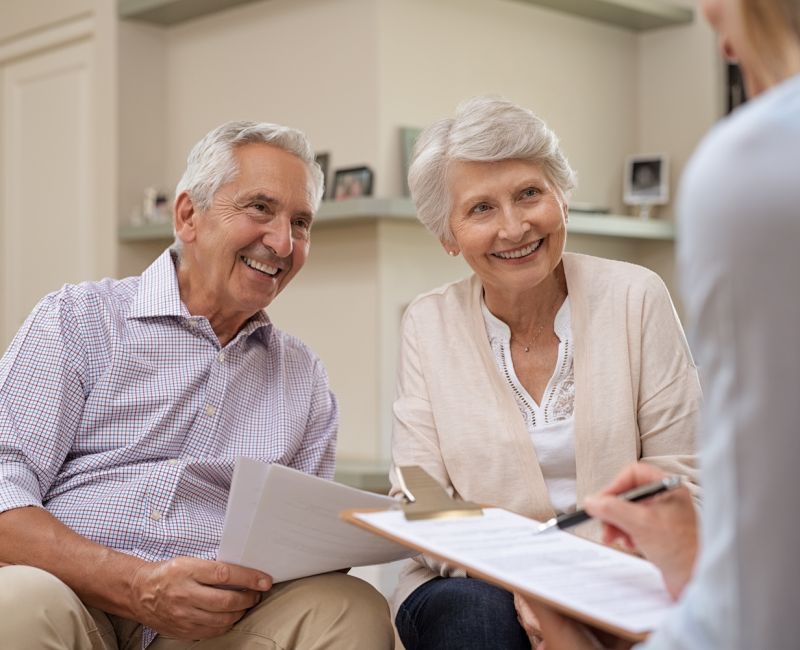 Senior couple discussing financial planning with advisor, smiling and holding documents in cozy living room setting.