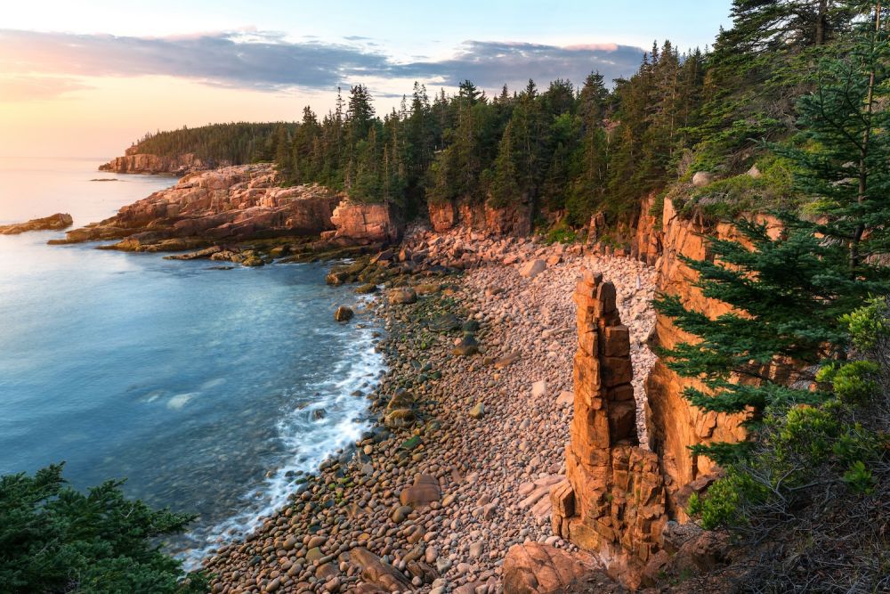 Rocky coastal landscape with cliffs and ocean waves at sunset, bordered by evergreen trees.