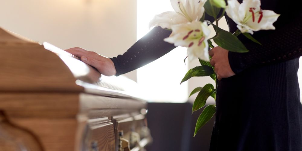 Person holding lilies, placing hand on wooden casket during funeral ceremony, expressing respect and condolences.