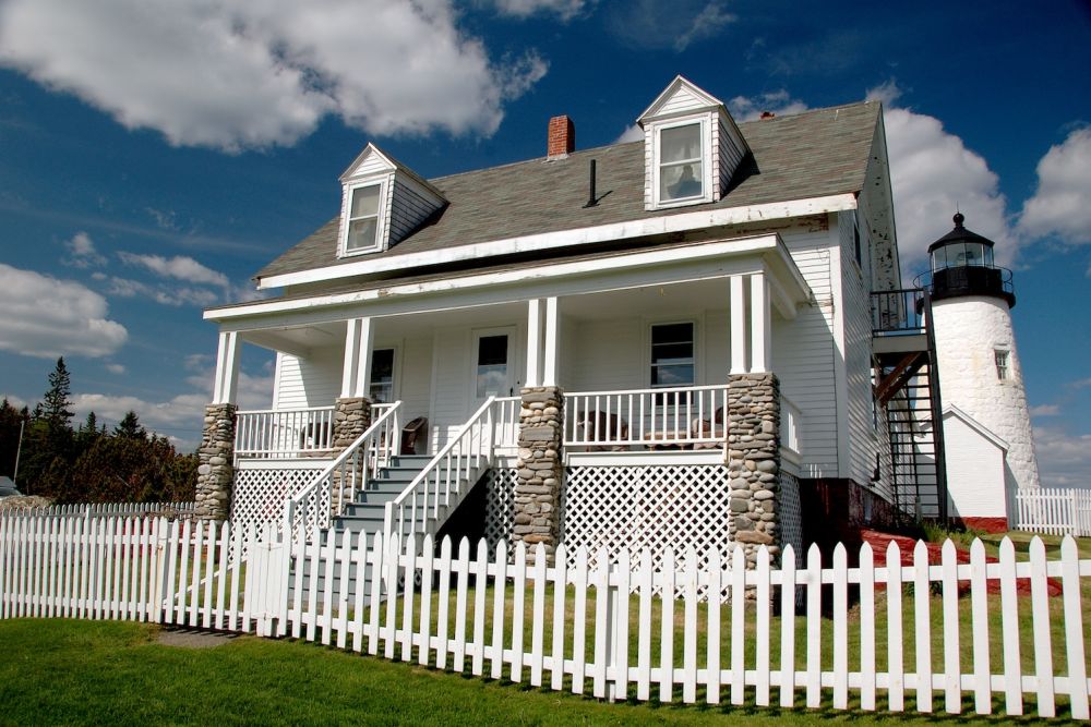 White house with stone pillars and picket fence near a lighthouse under a blue sky.